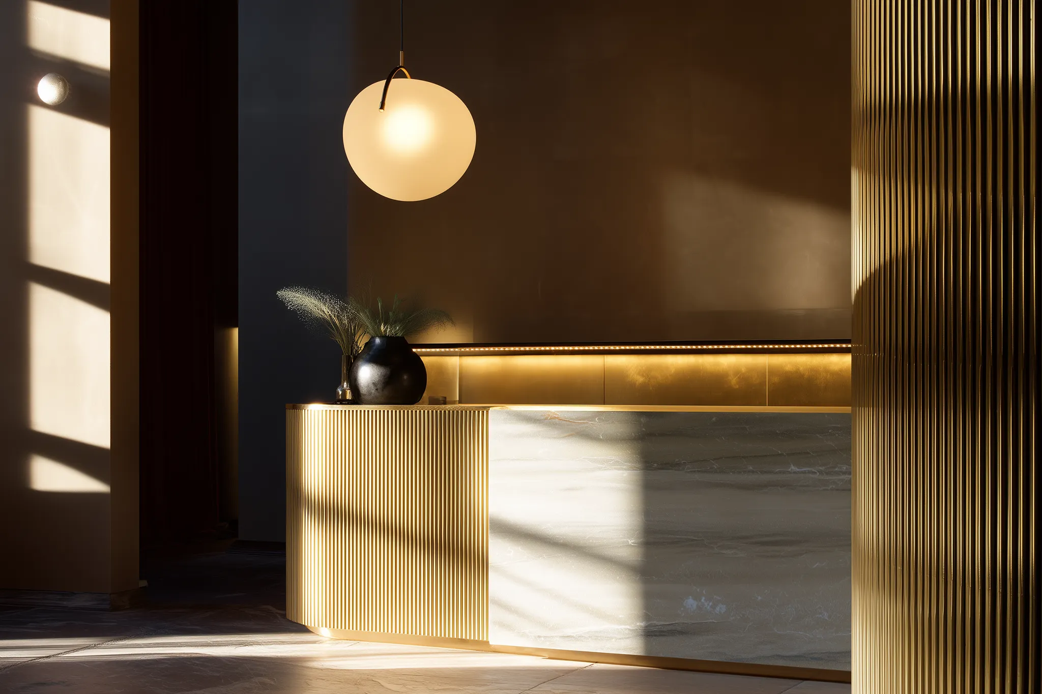 Modern reception desk with golden vertical slats, marble front panel, a round pendant light, and a black vase with decorative grass in soft natural light.